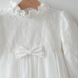 May include: White lace dress with a tulle skirt and a satin bow. The dress has long sleeves and a high neckline.