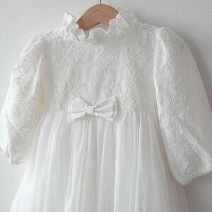 May include: White lace dress with a bow at the waist and long sleeves. The dress is made of a soft, delicate fabric and has a vintage look.