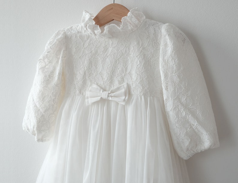 May include: White lace dress with a bow at the waist and long sleeves. The dress is made of a soft, delicate fabric and has a vintage look.