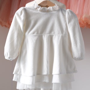 May include: White long-sleeved dress with a lace collar and a layered skirt.