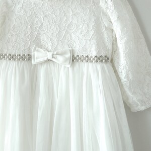 May include: White lace baby dress with a tulle skirt and a satin bow. The dress has a beaded belt with a pearl-like design.