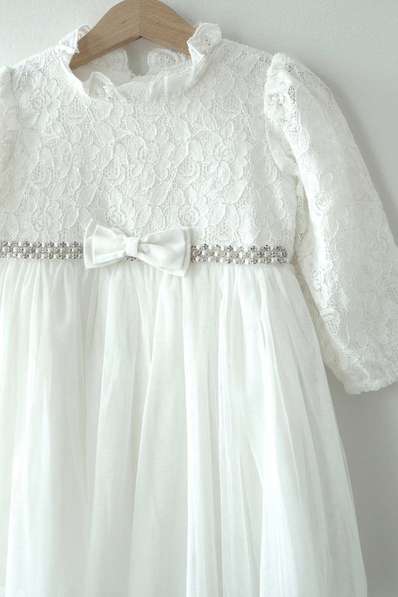 May include: White lace baby dress with a tulle skirt and a satin bow. The dress has a beaded belt with a pearl-like design.