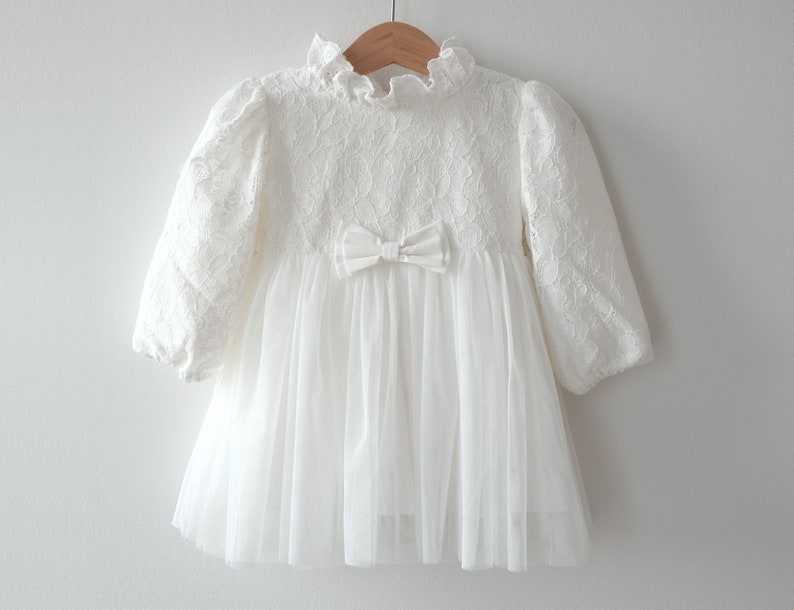 May include: White lace dress with a tulle skirt and a satin bow at the waist.