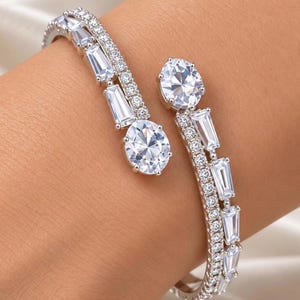 May include: A silver-toned open cuff bracelet featuring two oval-cut clear stones at the ends. The band is accented with a row of small round clear stones and a series of rectangular baguette-cut clear stones arranged in a linear pattern.