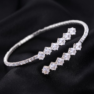 May include: A silver-toned bypass bracelet featuring a flexible band encrusted with small clear crystals. The two open ends of the bracelet are each adorned with a row of five square-cut clear stones, creating a sparkling, geometric design.