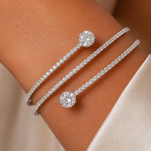 May include: A silver-toned bypass cuff bracelet featuring a continuous row of small pavé-set clear stones along the band. The bracelet ends in two larger, round, brilliant-cut clear stones that sit prominently at the open terminals of the design.