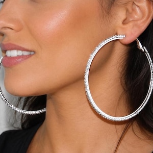 May include: Large silver-toned hoop earrings featuring a continuous row of small, clear, sparkling stones embedded along the outer surface. These circular earrings have a polished metallic finish and a secure hinged clasp mechanism for closure.