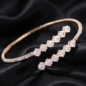 May include: A gold-toned open cuff bracelet featuring a flexible band encrusted with small clear crystals. The two ends of the bracelet terminate in parallel diagonal rows of larger, square-cut clear crystals that overlap slightly.
