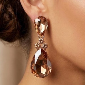 May include: A drop earring featuring a large, faceted, pear-shaped champagne-colored crystal suspended from a smaller, matching teardrop crystal. The two crystals are connected by a small, decorative metal setting with tiny, clear accent stones.