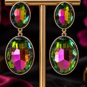 May include: A pair of drop earrings featuring two oval-shaped, multi-faceted iridescent crystals in each piece. The crystals display a vibrant spectrum of green, pink, and yellow hues, set within polished gold-tone metal frames and connected by small rings.