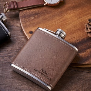Puede incluir: Una petaca de acero inoxidable cubierta de cuero marrón con una tapa plateada y el texto grabado "Niko GROOMSMAN".