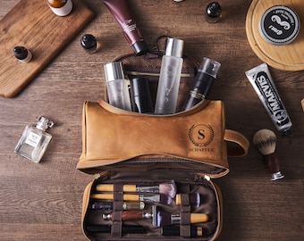 May include: A tan leather toiletry bag with a top handle and a personalized monogram featuring the letter S and the name SCHAFFER. The open bag reveals internal elastic loops holding various makeup brushes and a razor, with bottles and tubes nearby.
