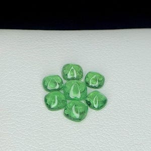 May include: Seven translucent, emerald-green gemstones arranged in a circular pattern. Each gem has a rounded, irregular shape, and they are displayed against a white background with a black backdrop.