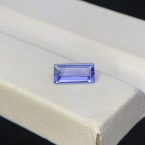 Puede incluir: Una gema azul rectangular y facetada. La gema se encuentra sobre una superficie blanca, con un fondo oscuro. Las facetas de la gema reflejan la luz, creando un efecto brillante. La gema es de un tono azul vibrante.