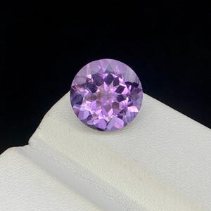 May include: A round, faceted amethyst gemstone. The gemstone is a light purple color with a brilliant cut, showcasing multiple facets that reflect light. The gem is set against a white surface with a black background.