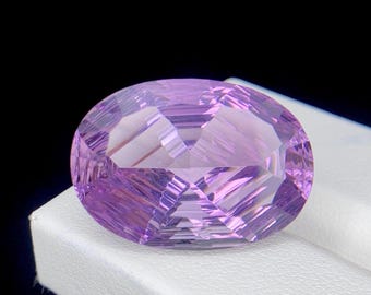 May include: A faceted, oval gemstone in shades of lavender and purple. The gem is set against a white surface with a black background. The cut of the stone creates a play of light and shadow, highlighting its facets.