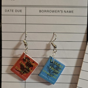 May include: Silver-tone dangle earrings featuring miniature book charms. One charm is red with "A Court of Thorns and Roses" text, the other is blue with "A Court of Frost and Starlight" text. The author's name, Sarah J Maas, is also visible.