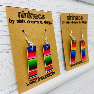 May include: Two pairs of rectangular earrings with a colorful striped pattern, displayed on brown cards. The earrings have silver-colored hooks and the cards feature the text "ninimacs by nini's dreams & things" and a website address.