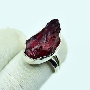 Op de afbeelding: Een zilveren ring met een grote, ruw gesneden, dieprode edelsteen. De edelsteen heeft een ruw, getextureerd oppervlak en is gezet in een gepolijste zilveren band. Het ontwerp van de ring is gedurfd en opvallend.