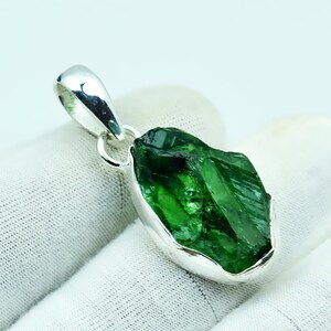 May include: A vivid green gemstone pendant with a silver-coloured bail. The raw, uncut stone is set in a polished silver setting, highlighting its natural, irregular form. The pendant is shown against a white backdrop.