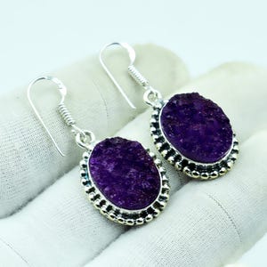 May include: A pair of silver earrings featuring oval, deep purple gemstone pendants. The gemstones have a textured, crystalline appearance and are set within a beaded silver frame. The earrings have a hook closure.
