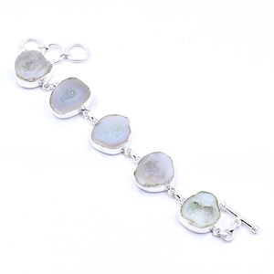 May include: A silver bracelet featuring five oval, light-colored geode stones. The stones are connected by silver links and have a light-colored interior. The bracelet has a toggle clasp.