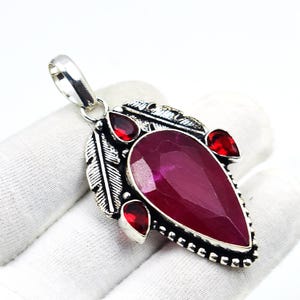 May include: A silver pendant with a large, pear-shaped, deep red gemstone as the focal point. The pendant is adorned with smaller red gemstones and decorative silver feather accents. The bail is silver, and the pendant is set against a white background.