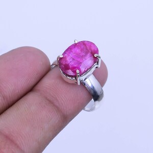 May include: A silver ring featuring a large, oval-cut, hot pink gemstone. The gemstone is set in a silver band with four prongs. The ring is held up by a finger.