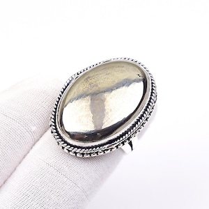 May include: An oval-shaped ring featuring a large, smooth, metallic pyrite stone set in a silver-toned metal frame. The setting is detailed with a decorative twisted rope border and a beaded edge, creating a textured appearance around the stone.