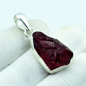 Pode incluir: Um pingente de prata com uma pedra preciosa vermelha escura bruta. A pedra de formato irregular é colocada em uma moldura de prata polida com uma fiança combinando para um colar. A pedra preciosa tem uma superfície áspera e texturizada.