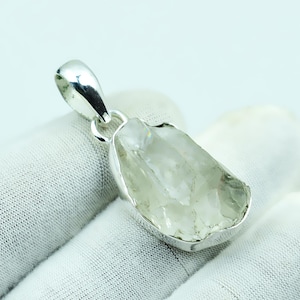 May include: A clear, raw quartz pendant with a silver bezel and bail. The quartz has a rough, natural shape and is set in a polished metal setting. The pendant is displayed on a white surface.