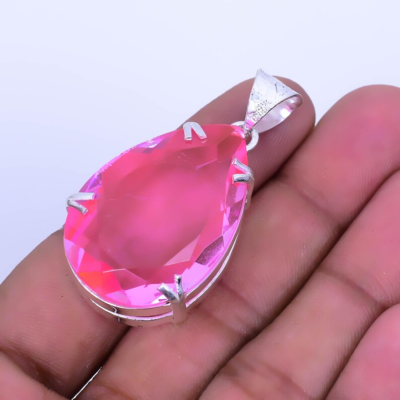 May include: A pink pear-shaped gemstone pendant with a silver setting. The pendant is held in a hand.