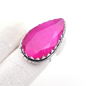 Può includere: Un anello a forma di goccia con una grande pietra preziosa sfaccettata di colore rosa vibrante, incastonata in una lunetta di metallo color argento. L'incastonatura presenta un bordo decorativo dentellato che incornicia la pietra testurizzata.