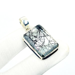 Pode incluir: Pingente retangular com uma pedra clara de cor clara com inclusões lineares pretas. A pedra é engastada em uma moldura de metal prateado com uma fiança combinando para um colar. O pingente é exibido em um fundo branco.