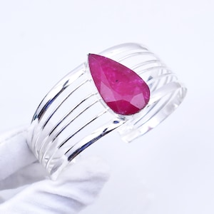 May include: A silver cuff bracelet with a large, teardrop-shaped, faceted ruby gemstone. The bracelet has a multi-band design with a polished finish. The ruby is a vibrant shade of pink.