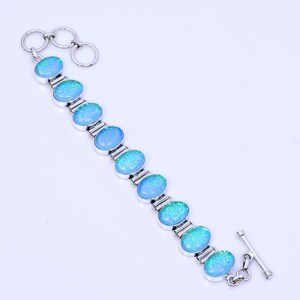 Puede incluir: Un brazalete de plata con un diseño de cadena de eslabones y piedras ovaladas de ópalo azul. El brazalete tiene un cierre de palanca.
