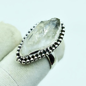 May include: A silver ring with a large, clear quartz crystal. The quartz is an elongated, irregular shape, set in a silver bezel with a beaded border. The ring's band is also silver.