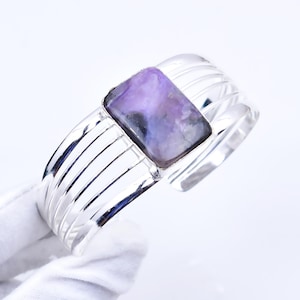 Pode incluir: Uma pulseira de punho prateada com uma pedra preciosa retangular roxa. A pulseira tem um design canelado e um acabamento polido. A pedra preciosa exibe uma mistura de tons roxos e pretos.