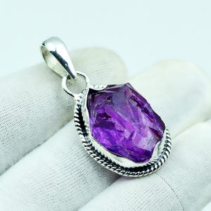 May include: A silver pendant with a raw, faceted amethyst gemstone. The purple crystal is set in a silver bezel with a twisted rope design around the edge. The pendant has a silver bail for a chain.