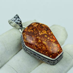 Puede incluir: Un colgante rectangular de ámbar con una fianza decorativa de color plateado. El ámbar tiene un color marrón cálido y rico con motas doradas. El colgante está enmarcado con un borde plateado detallado.