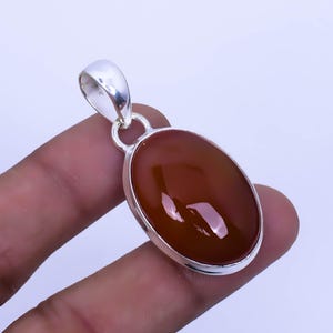 Pode incluir: Um pingente de prata com uma pedra de cornalina oval e lisa. A cornalina é de cor vermelho-alaranjado escuro e está engastada em uma moldura de prata.