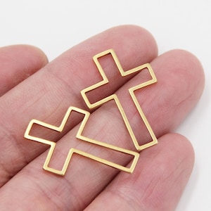 Puede incluir: Dos formas de cruz de metal de color dorado. La cruz más grande está ligeramente inclinada y la cruz más pequeña está directamente debajo.