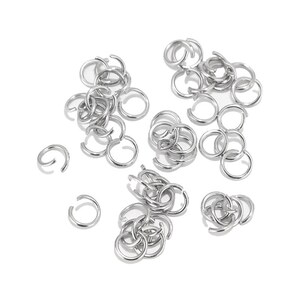 May include: A large quantity of silver jump rings, a type of jewellery finding, are scattered on a white background. The rings are small and circular, with a gap in the circle.