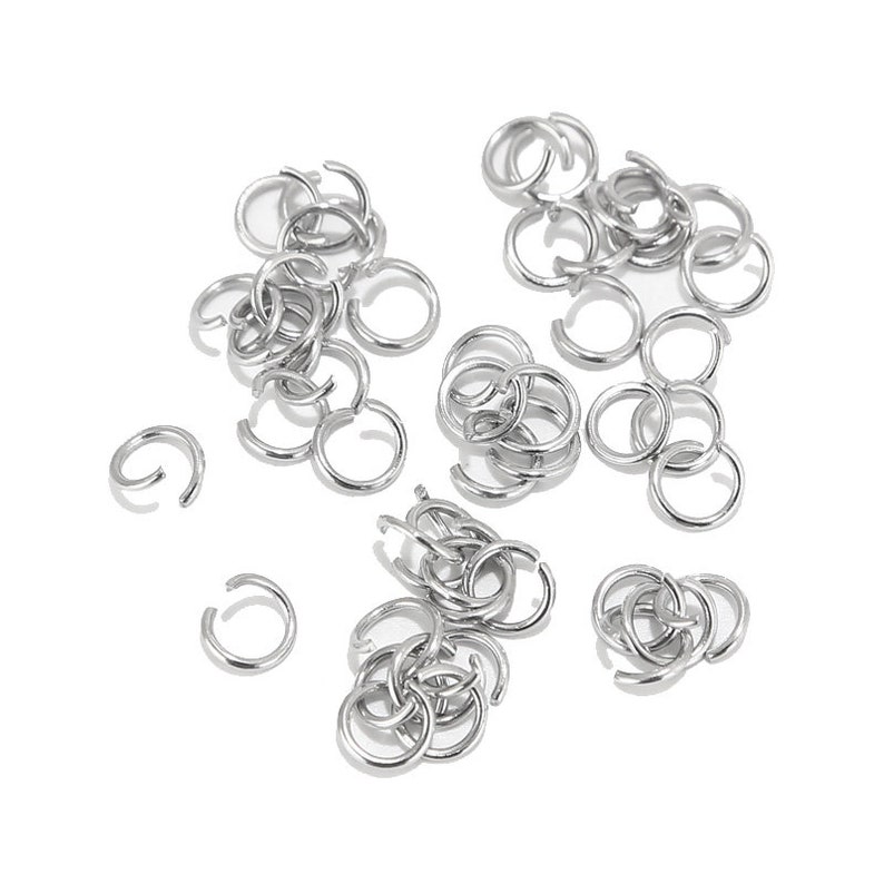 May include: A large quantity of silver jump rings, a type of jewellery finding, are scattered on a white background. The rings are small and circular, with a gap in the circle.