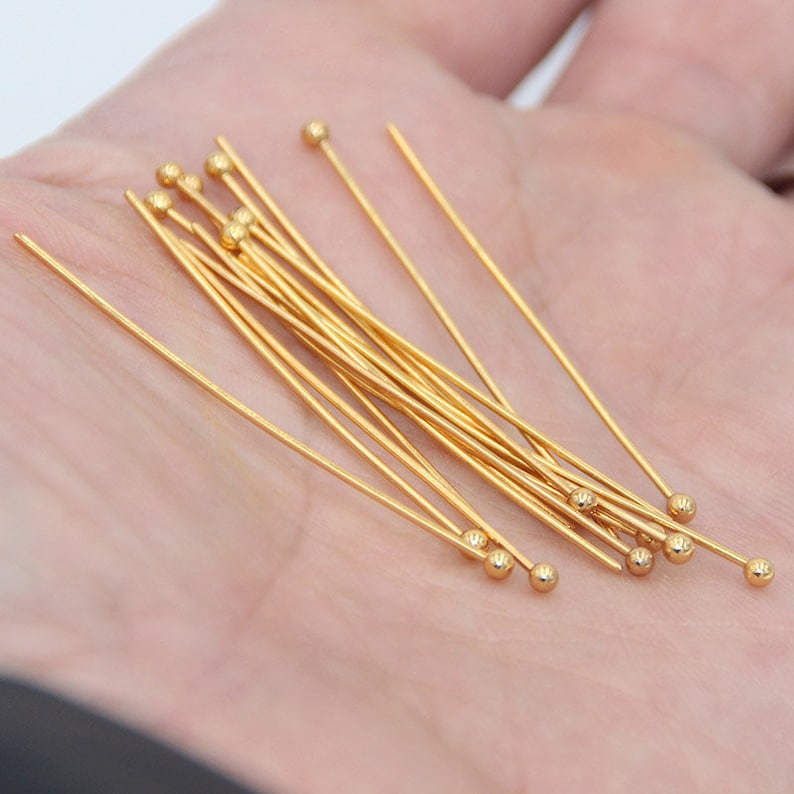 May include: A close-up of a pile of gold-colored head pins with round heads. The pins are arranged in a fan shape on a hand.