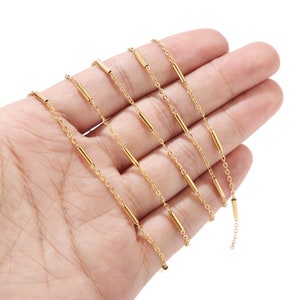 May include: A set of five gold-toned chain necklaces with small, cylindrical beads. The chains are layered on top of each other and are displayed on a hand.
