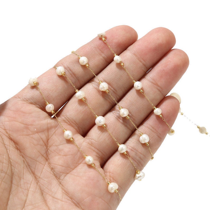 Puede incluir: Un collar de cadena de oro con cuentas de perlas blancas. El collar est&aacute; colocado sobre una mano, mostrando la longitud y el detalle de la cadena.