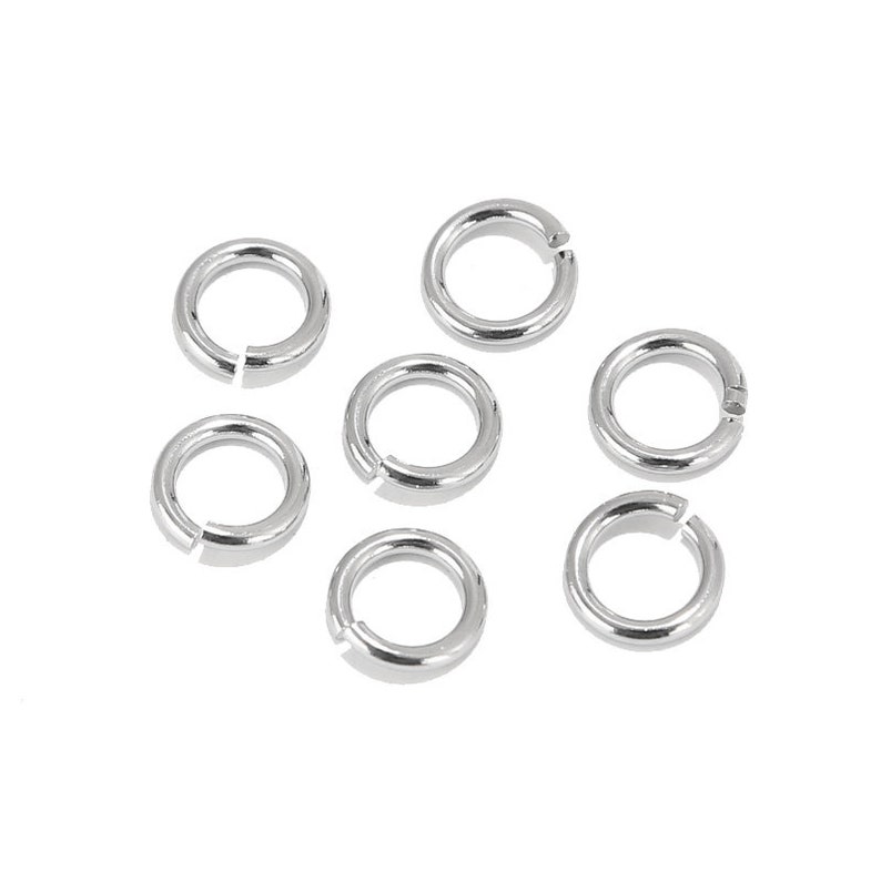 May include: Seven silver jump rings, also known as split rings, are arranged on a white surface. These rings are commonly used in jewellery making and crafting.