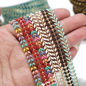 Könnte beinhalten: Nahaufnahme einer goldenen Kette mit bunten Emaille-Akzenten. Die Kette besteht aus kleinen, ovalen Gliedern, die in einem Chevron-Muster angeordnet sind. Die Emaille-Akzente sind in verschiedenen Farben, darunter Rot, Blau, Grün, Gelb und Pink.