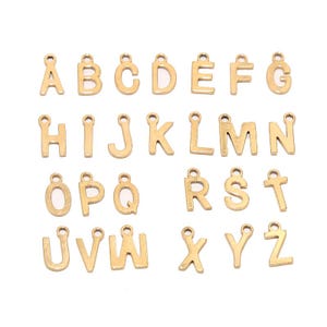 Könnte beinhalten: Goldfarbene Metall-Anhänger in Form von Buchstaben des Alphabets. Jeder Anhänger ist ein anderer Buchstabe des Alphabets, von A bis Z.